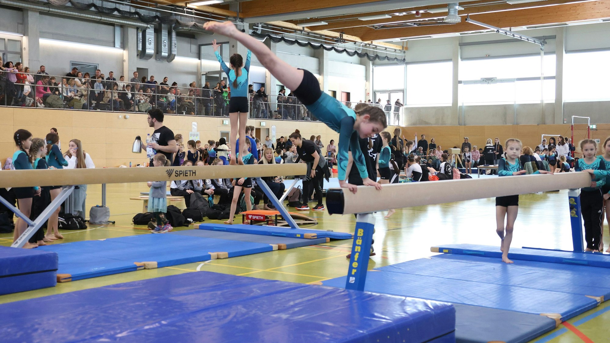 Ein Turnerin schwingt über den Balken, auf dem sie sich mit den Händen abstützt. Im Hintergrund ist eine volle Turnhalle zu erkennen.