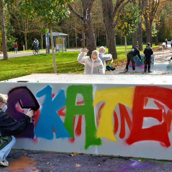 Jugendliche bringen mit Spraydosen Farbe auf die graue Skateanlage. Das Wort „Skate“ ist zu lesen, die Buchstaben in unterschiedlichen Farben.