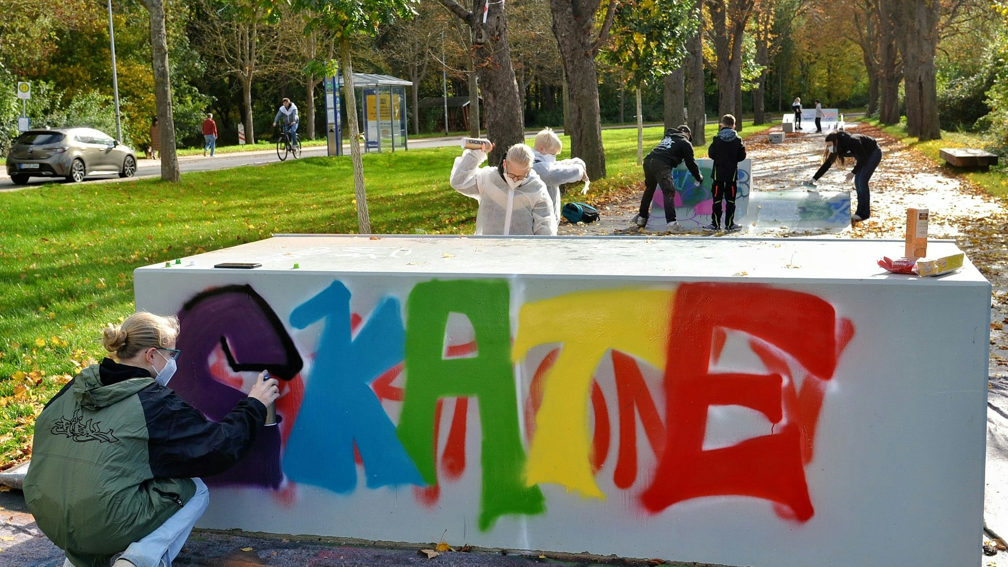 Jugendliche bringen mit Spraydosen Farbe auf die graue Skateanlage. Das Wort „Skate“ ist zu lesen, die Buchstaben in unterschiedlichen Farben.