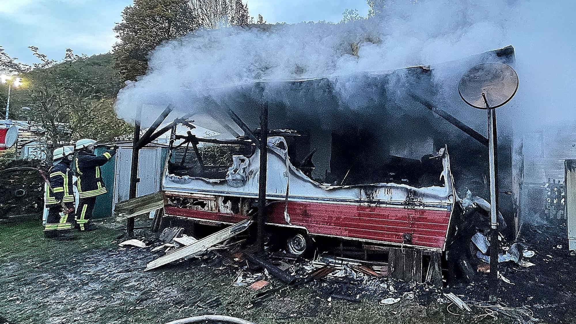 Zwei Feuerwehrleute stehen neben einem ausgebrannten Wohnwagen und einem qualmenden Überbau aus Holz.
