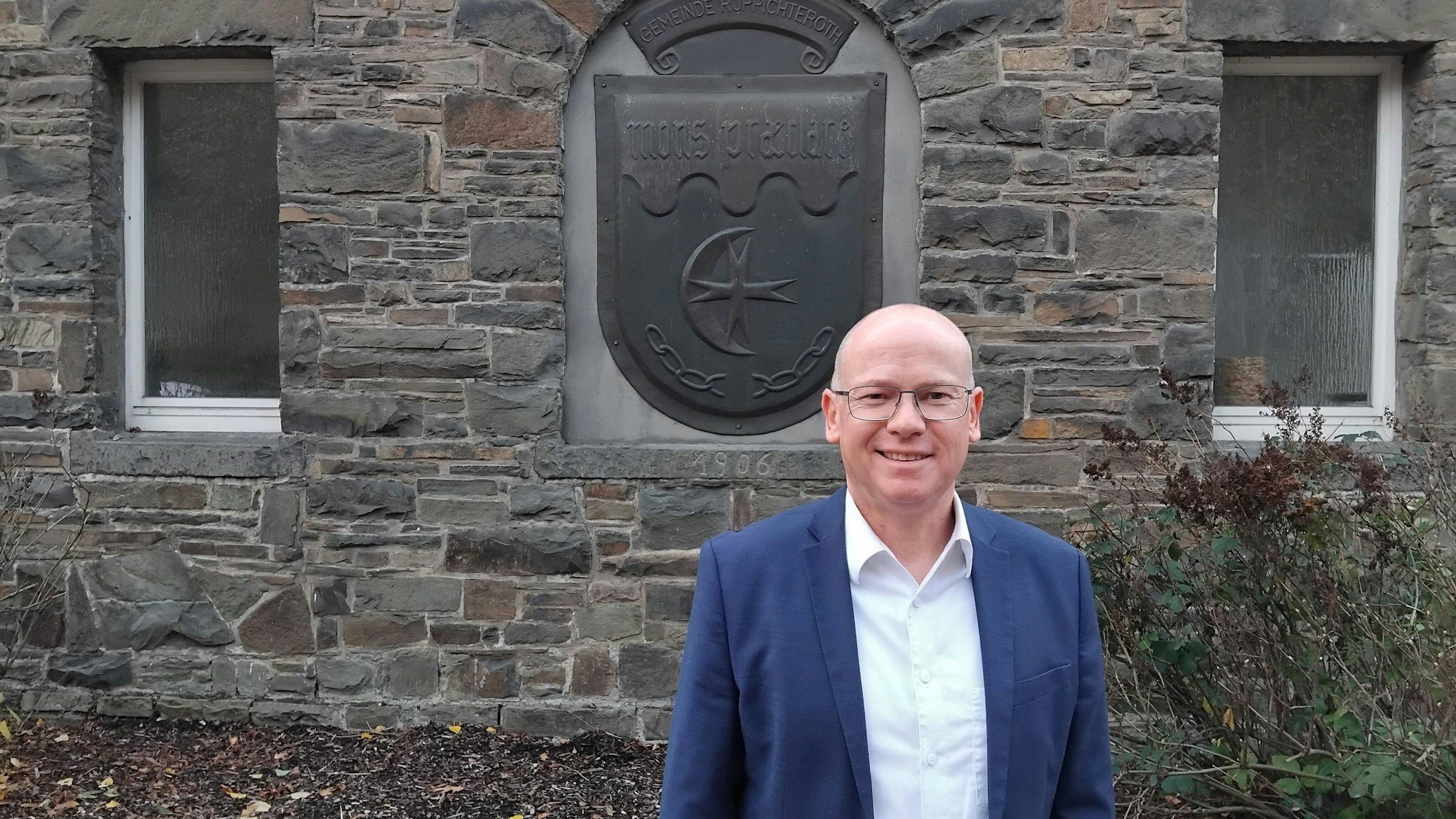 Mario Loskill, Bürgermeister von Ruppichteroth, vor dem Wappen am Rathaus Schönenberg, mit der lateinischen Aufschrift Mons Praeclare (ganz klarer Berg), die für Schönenberg steht.
