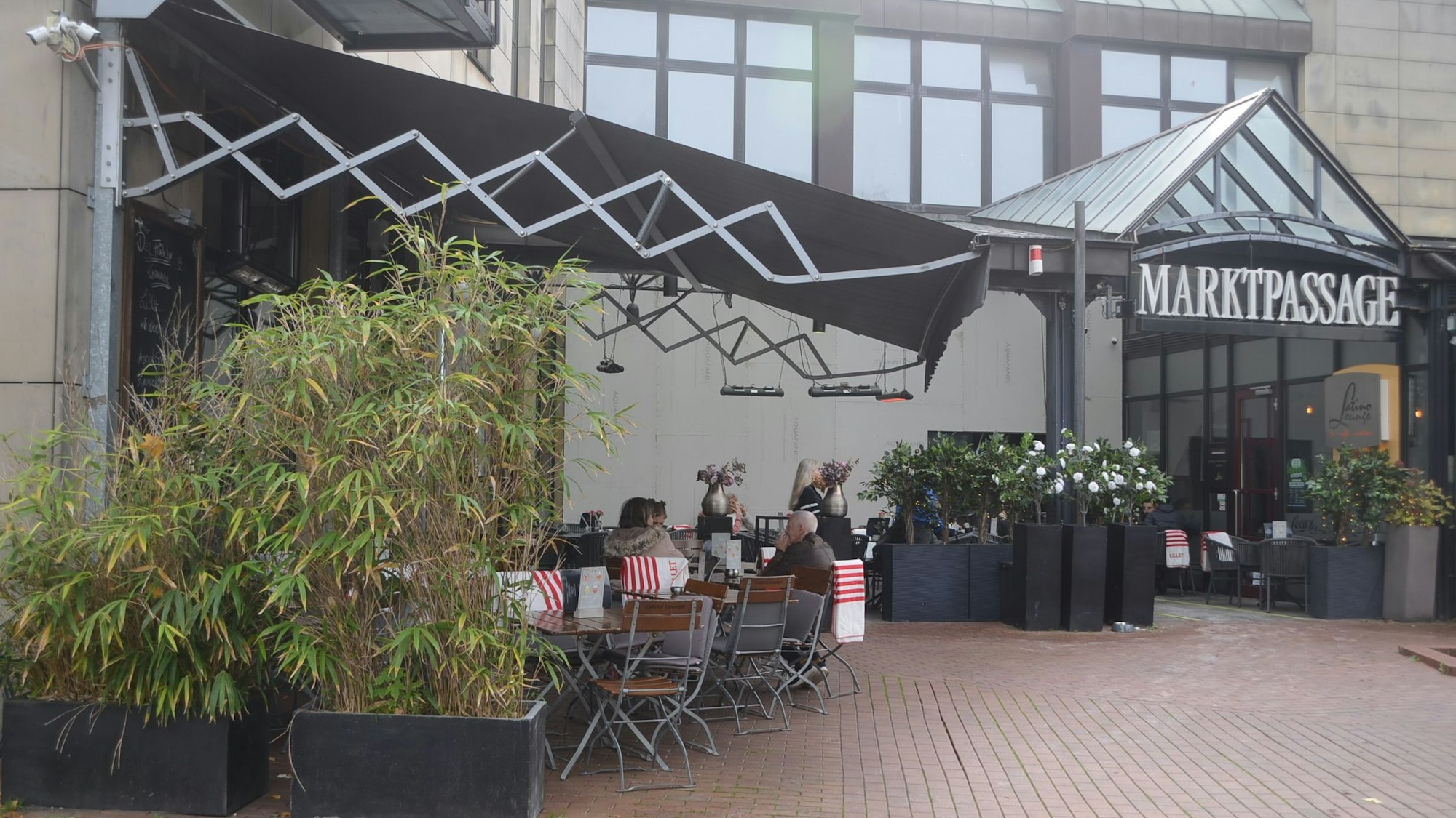 Tische und Markise einer Außengastronomie; unter einem Glasdach hängt der Schriftzug Marktpassage
