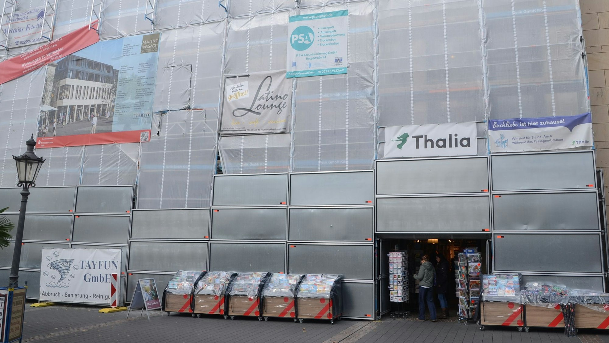 Netze und Metallbleche verhüllen eine Fassade. Davor stehen Büchertische, an der Fassade hängen Firmenschilder.