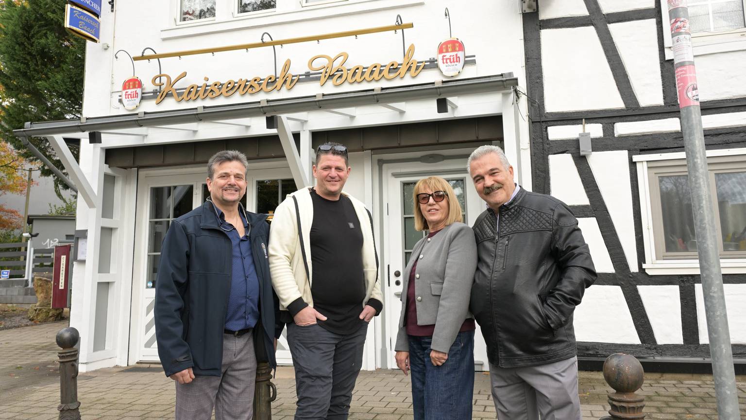 Drei Männer und eine Frau stehen vor einem Lokal mit der Aufschrift „Kaisersch Baach“.