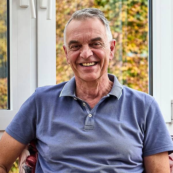 Rudolph Westerburg sitzt in einem blauen T-Shirt vor einem Fenster und lächelt in die Kamera.