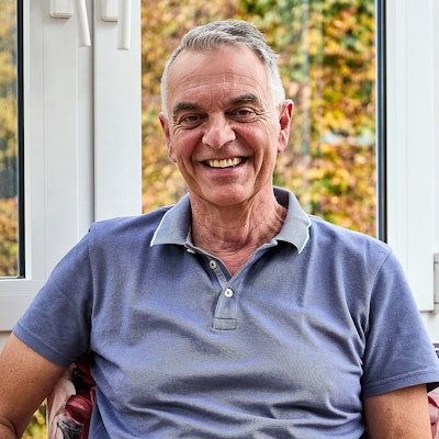 Rudolph Westerburg sitzt in einem blauen T-Shirt vor einem Fenster und lächelt in die Kamera.