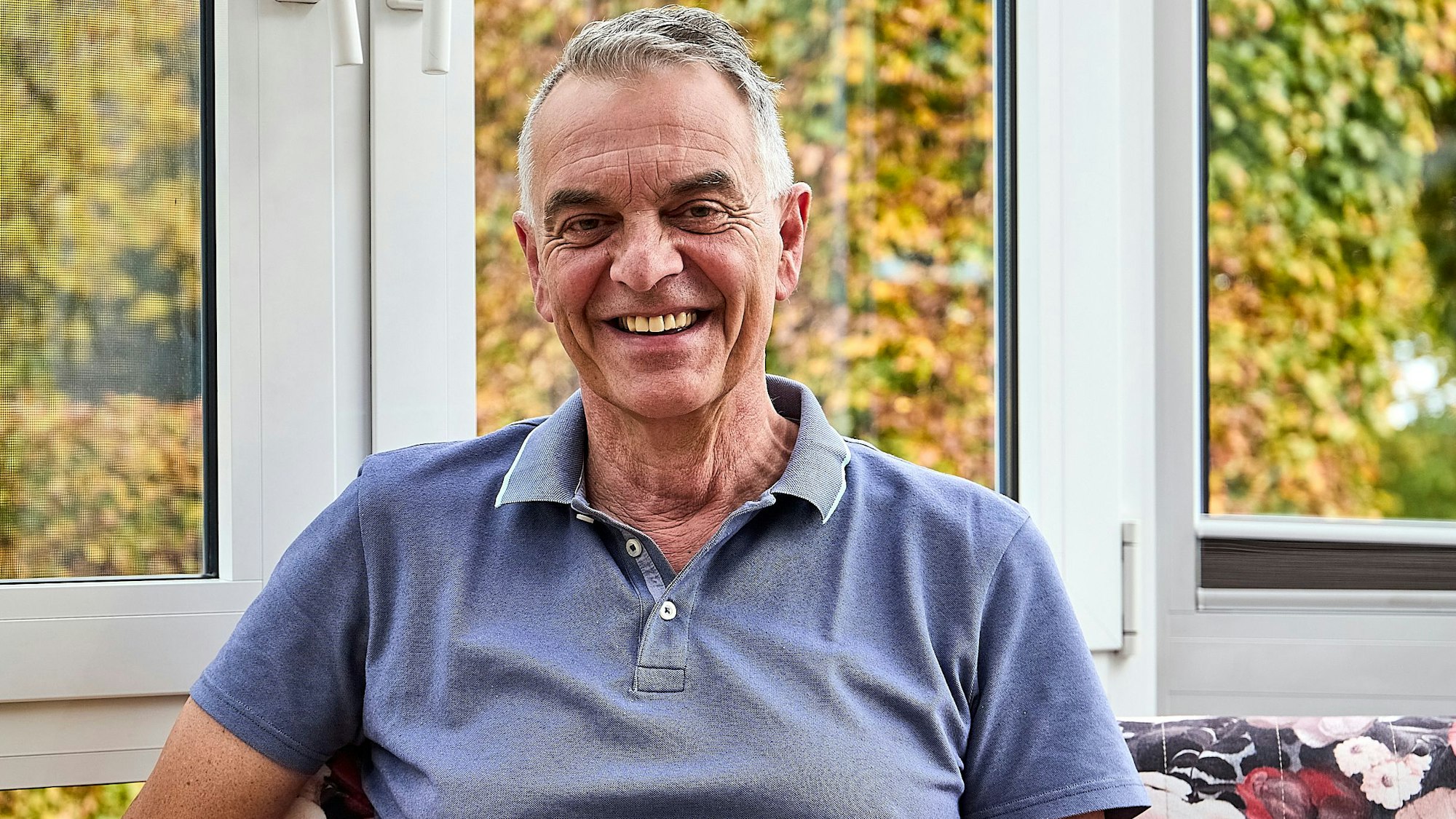 Rudolph Westerburg sitzt in einem blauen T-Shirt vor einem Fenster und lächelt in die Kamera.