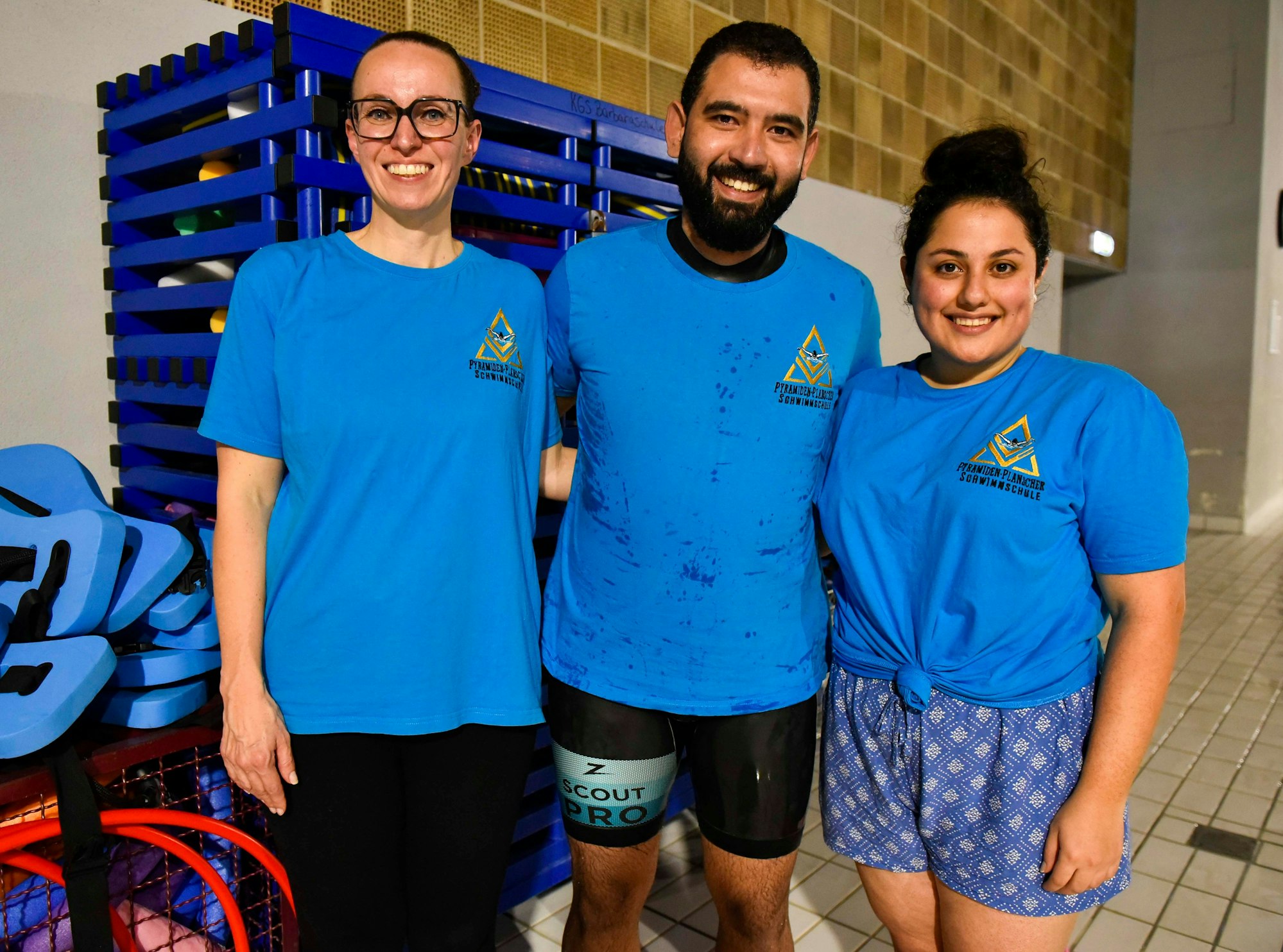 Alle drei tragen blaue T-Shirts. Im Hintergrund das Bad und Schränke mit Utensilien zum Schwimmen.