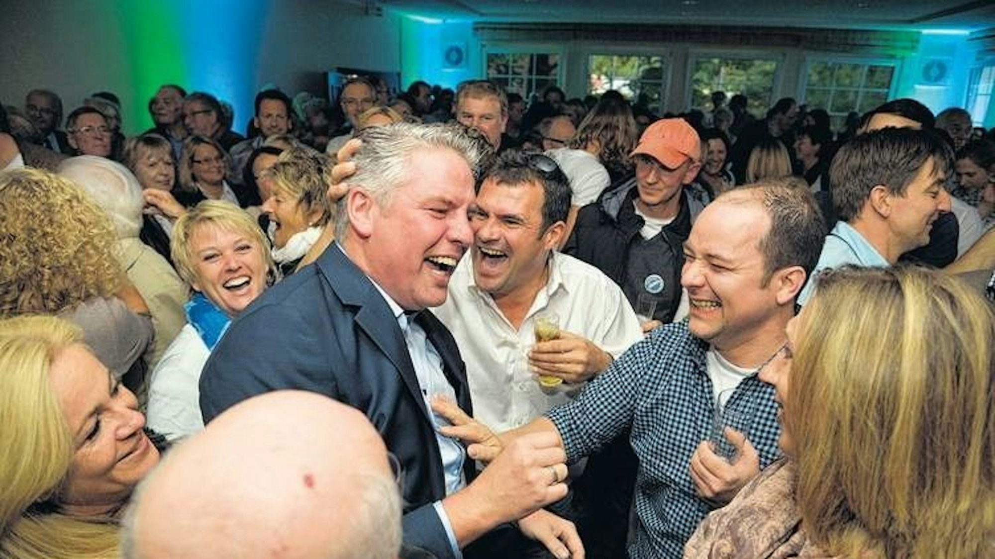 Eine Schar von Menschen bejubelt einen Mann in einem Anzug, der in einem Saal steht und lacht