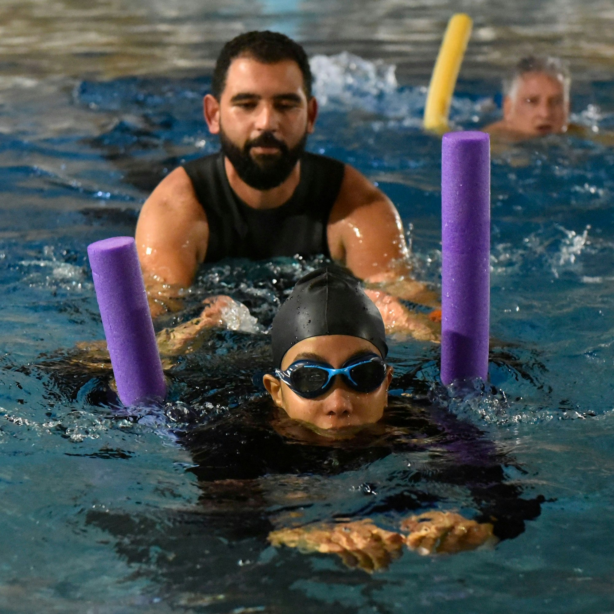 Zu sehen sind ein Schwimmer mit einer Schwimmnudel, Badekappe und Schwimmbrille. Hinter ihm ein Schwimmlehrer, der ihm im Wasser Unterstützung gibt.