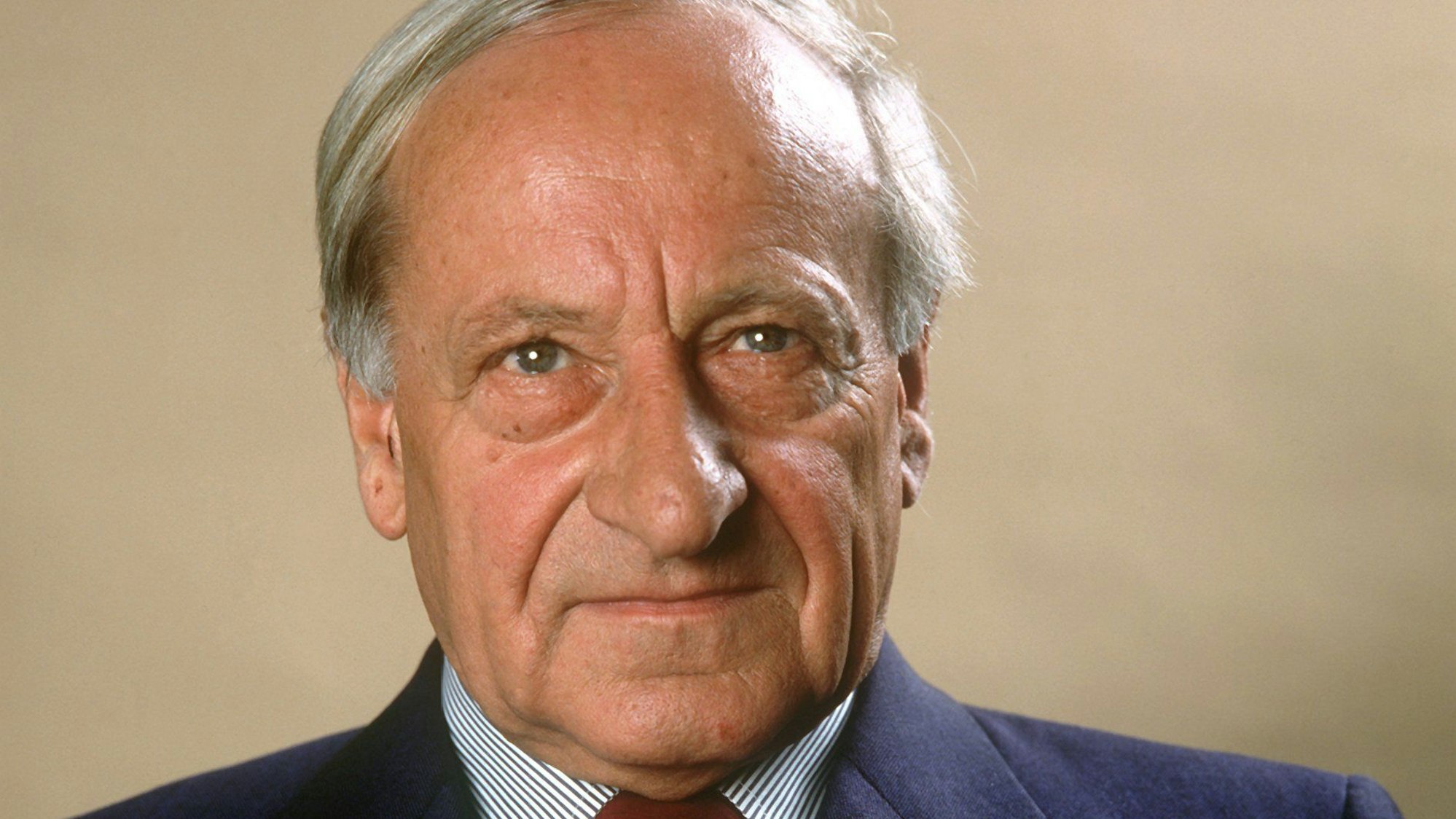 Auf dem Foto ist ein älterer Mann mit grauem Haar zu sehen. Er trägt ein blaues Jackett und eine rote Krawatte.