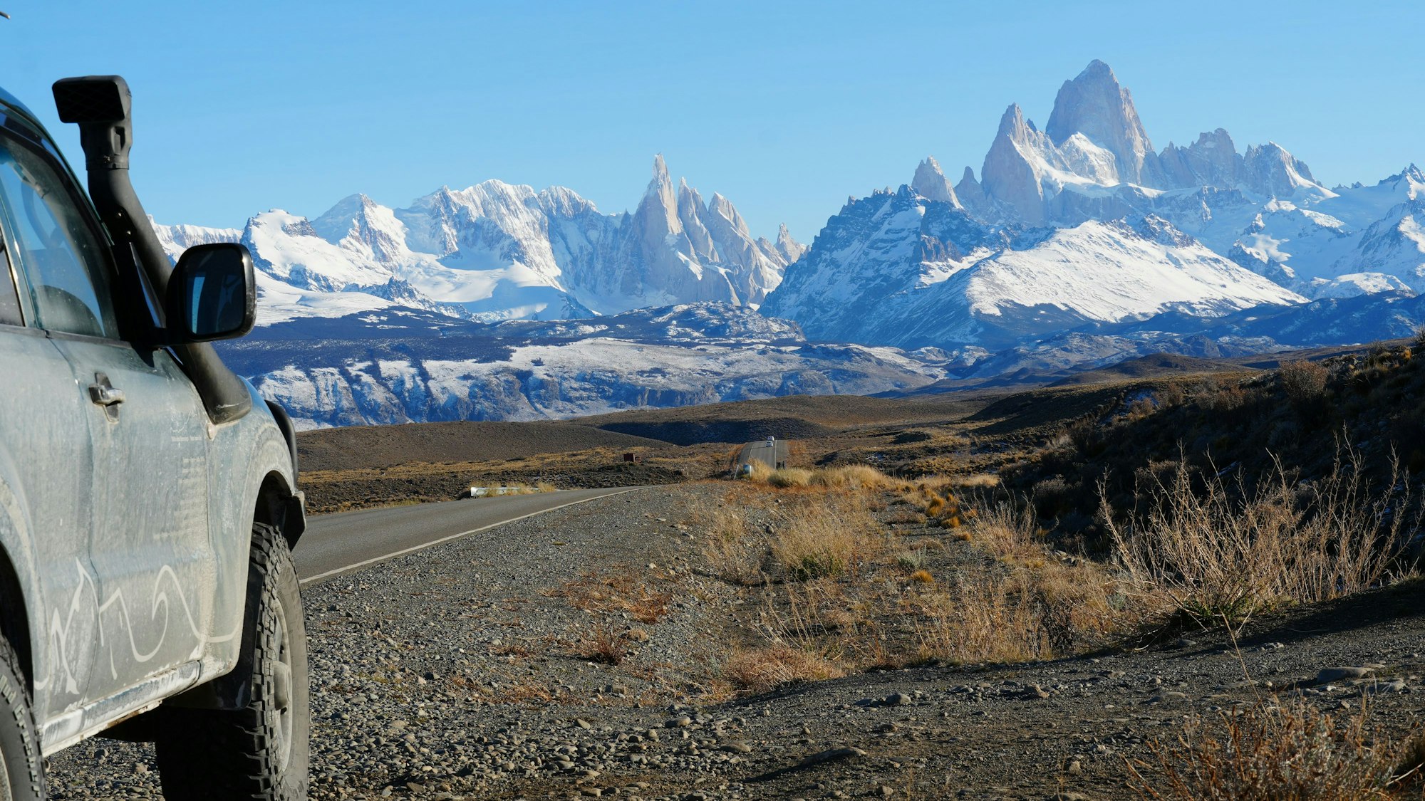 Blick auf eine bizarr geformte Gebirgskette der Anden. Am linken Bildrand steht ein Geländewagen.