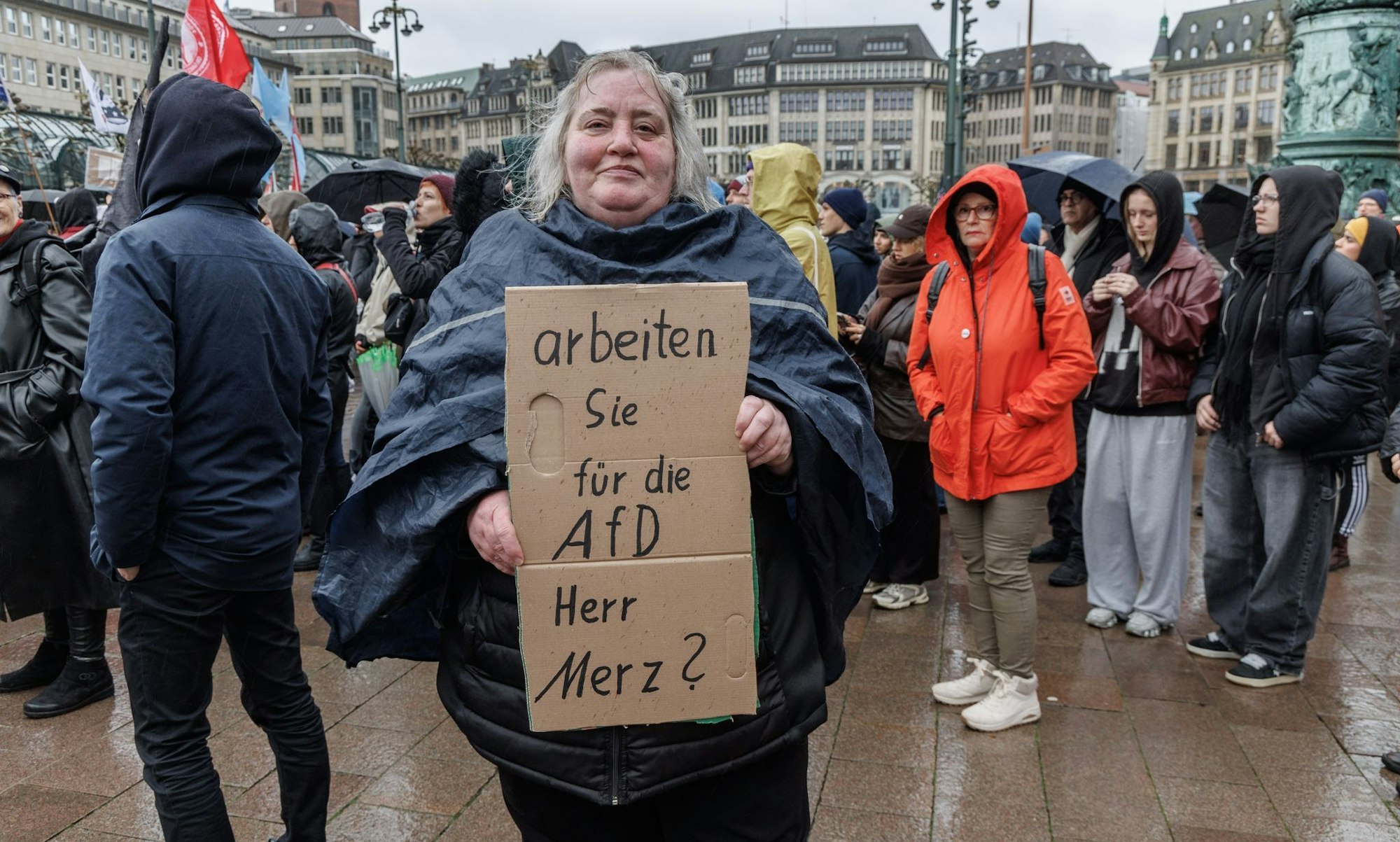 Eine Demonstrantin in Hamburg stellt auf einem Pappschild eine Frage an Bundeskanzler Friedrich Merz (CDU).