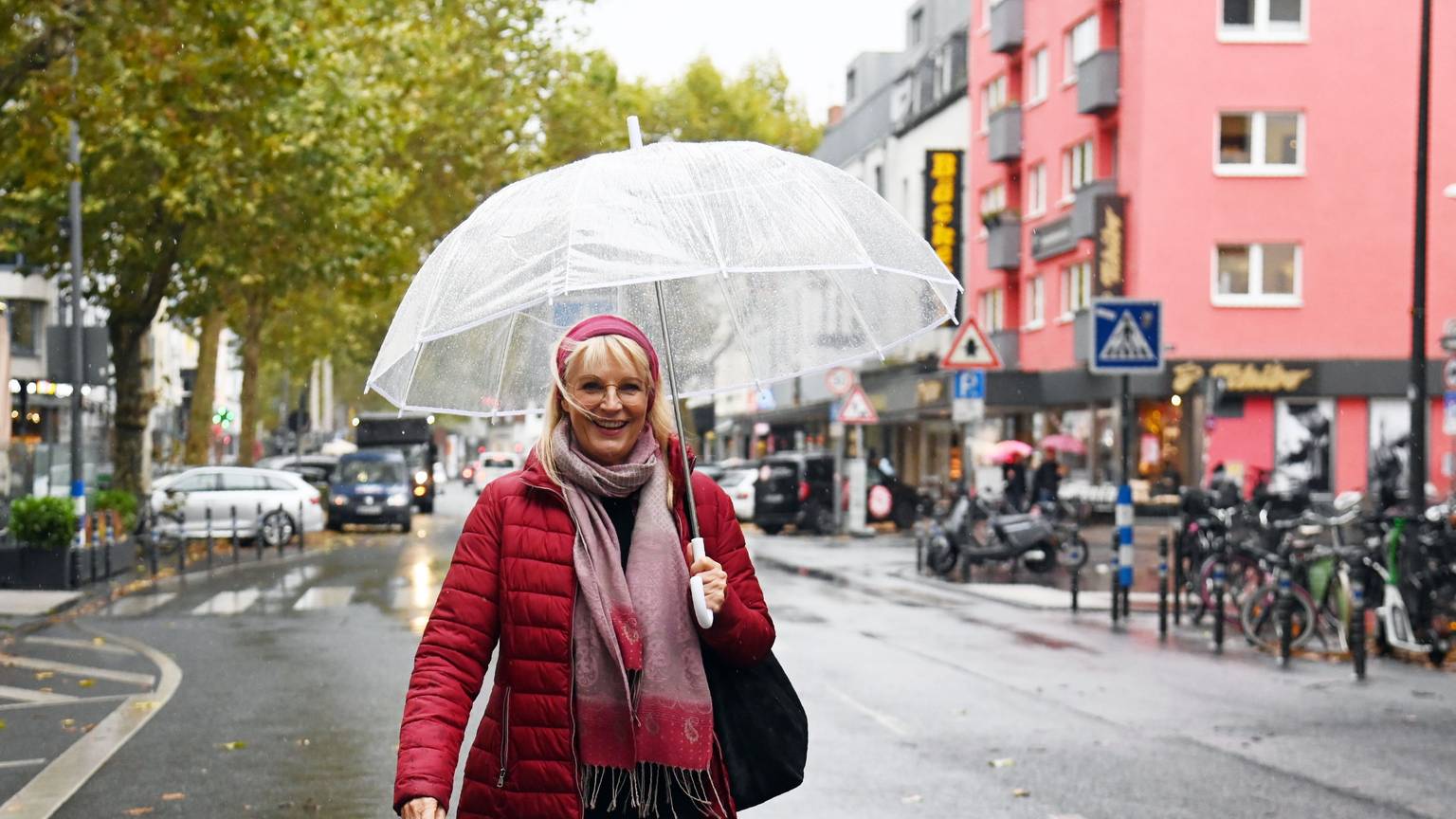 Eine Frau mit roter Jacke, rotem Haarband und durchsichtigem Regenschirm steht auf einer Geschäftsstraße.
