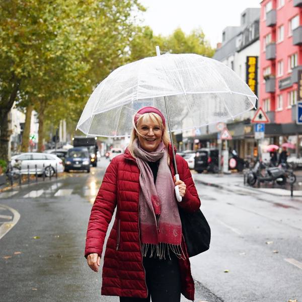 Eine Frau mit roter Jacke, rotem Haarband und durchsichtigem Regenschirm steht auf einer Geschäftsstraße.