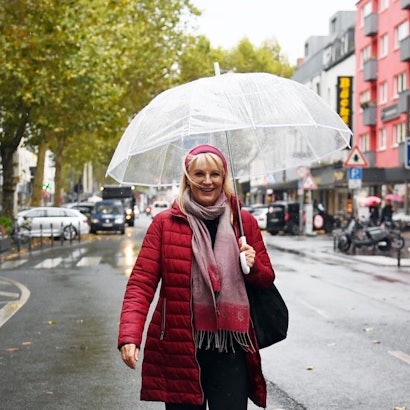 Eine Frau mit roter Jacke, rotem Haarband und durchsichtigem Regenschirm steht auf einer Geschäftsstraße.