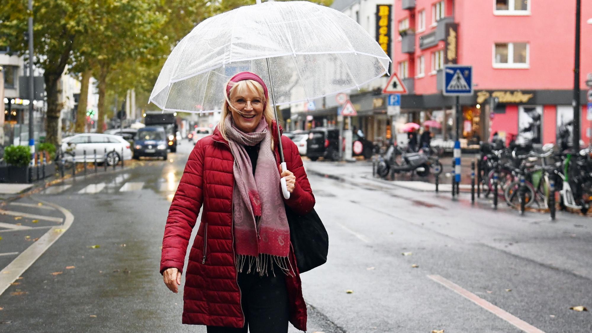 Eine Frau mit roter Jacke, rotem Haarband und durchsichtigem Regenschirm steht auf einer Geschäftsstraße.