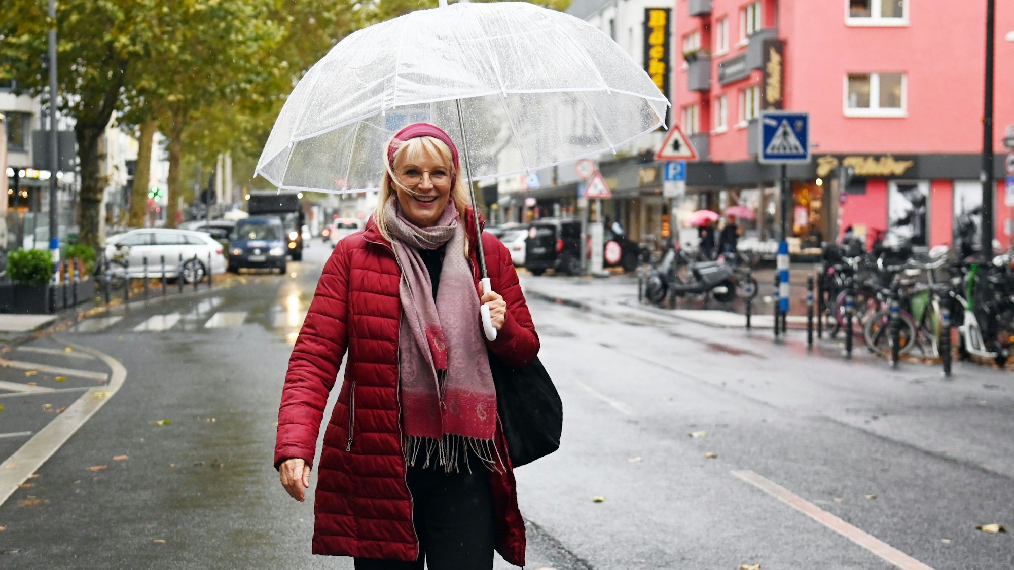 Eine Frau mit roter Jacke, rotem Haarband und durchsichtigem Regenschirm steht auf einer Geschäftsstraße.