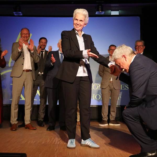 Ein Mann kniet vor einer Frau, im Hintergrund applaudieren mehrere Menschen.