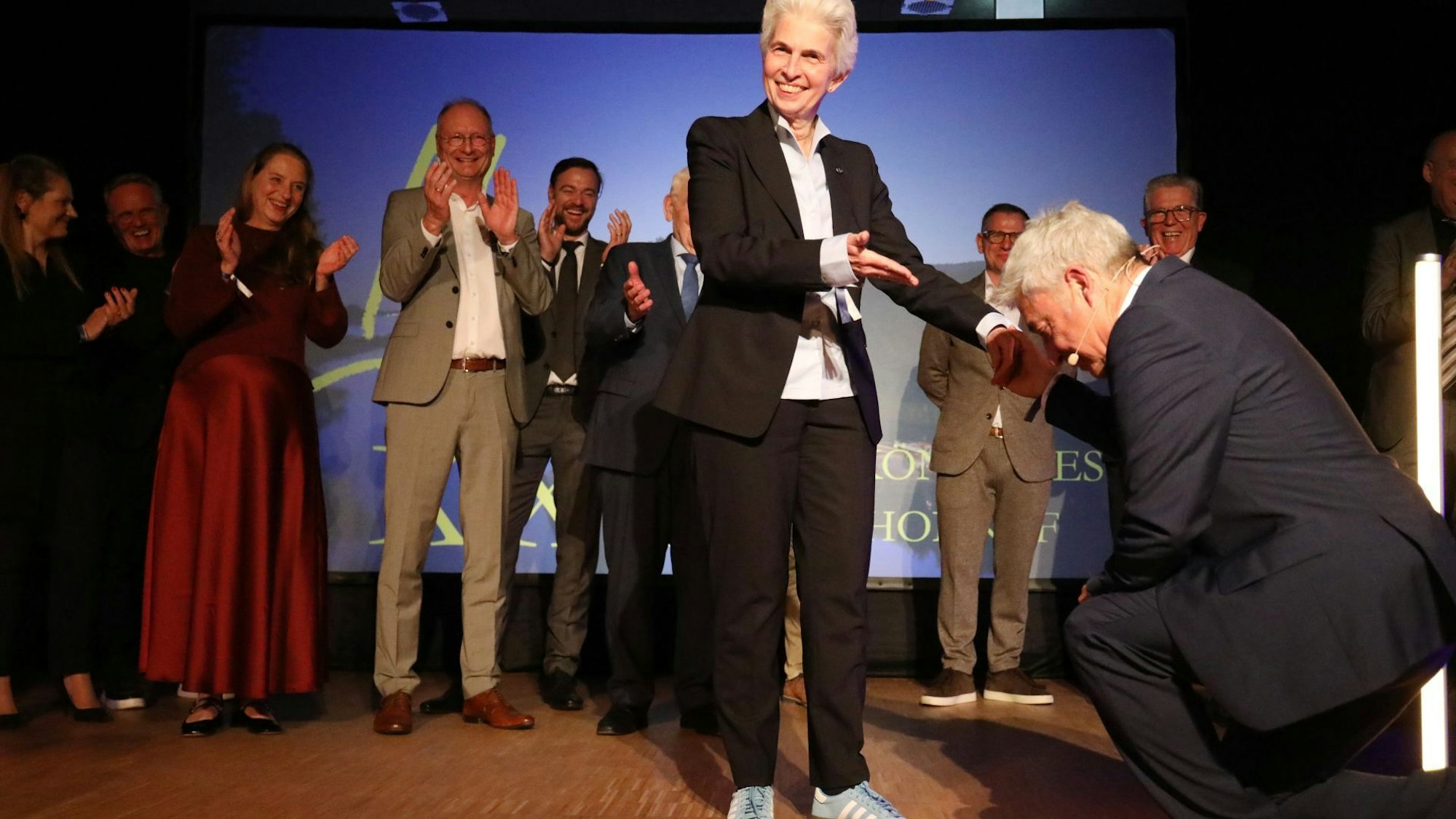 Ein Mann kniet vor einer Frau, im Hintergrund applaudieren mehrere Menschen.