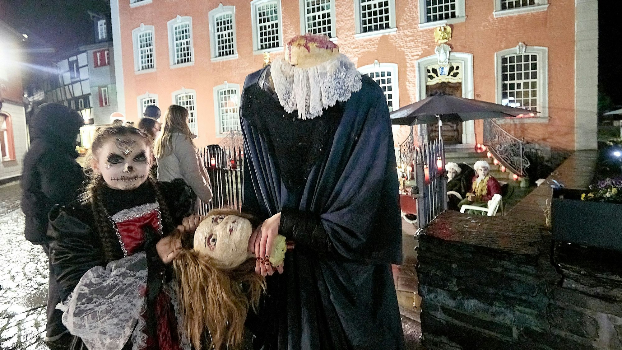 Zwei Figuren stehen vor dem Roten Haus in Monschau und halten einen Kopf in der Hand, der der einen Figur fehlt.