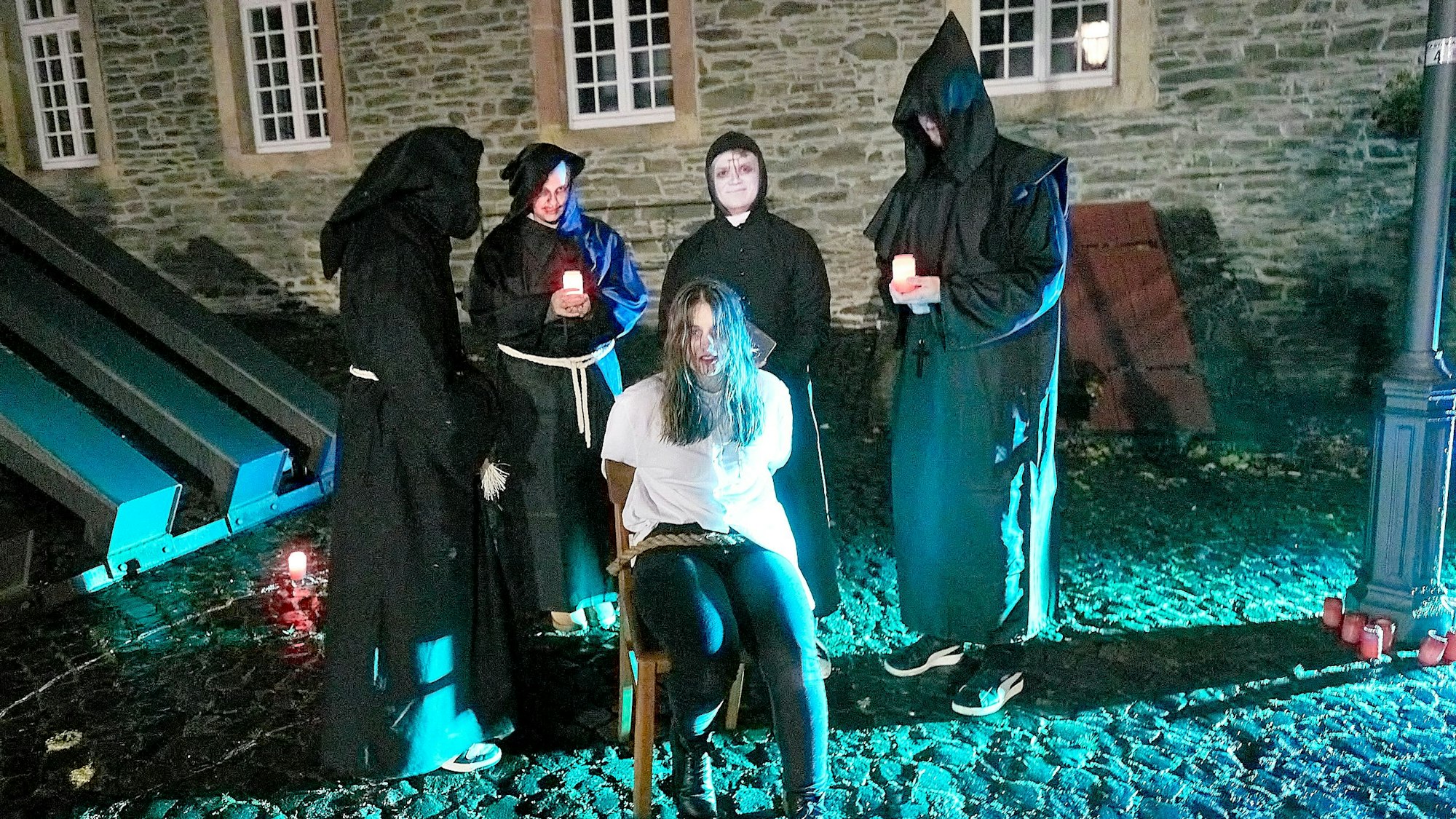 Mehrere Personen stehen in dunklen Roben um eine Frau herum, die auf einem Stuhl sitzt und festgebunden ist.