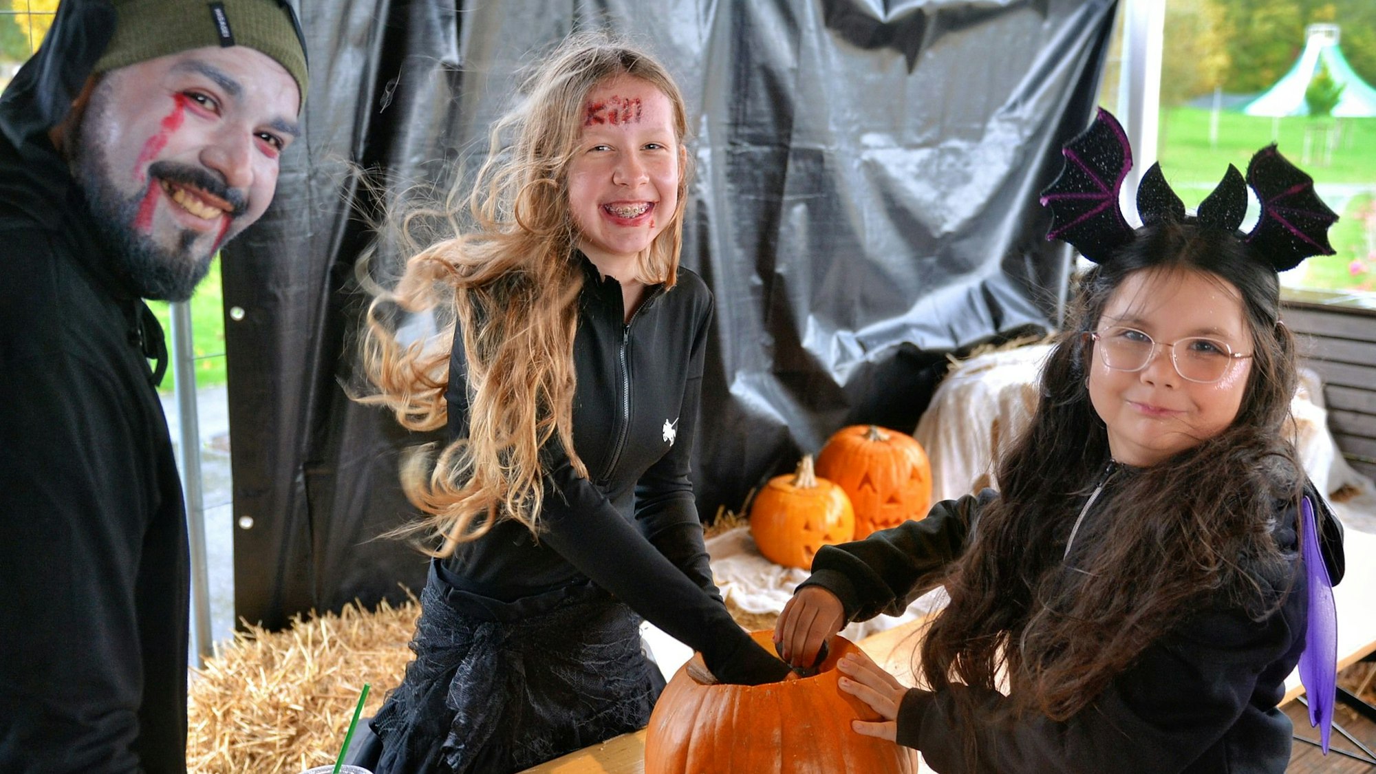 Talia, Charlyn und Vater Cihan (v.r.) stehen in schwarzen Kostümen an einem Tisch und schnitzen Kürbisse.