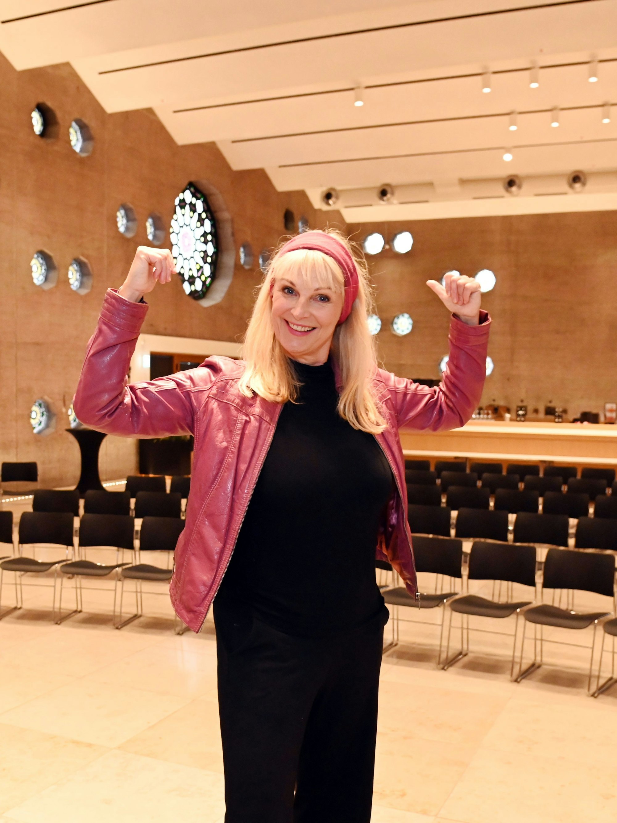 Eine Frau mit roter Lederjacke steht in einem großen Veranstaltungsraum in Siegerpose.
