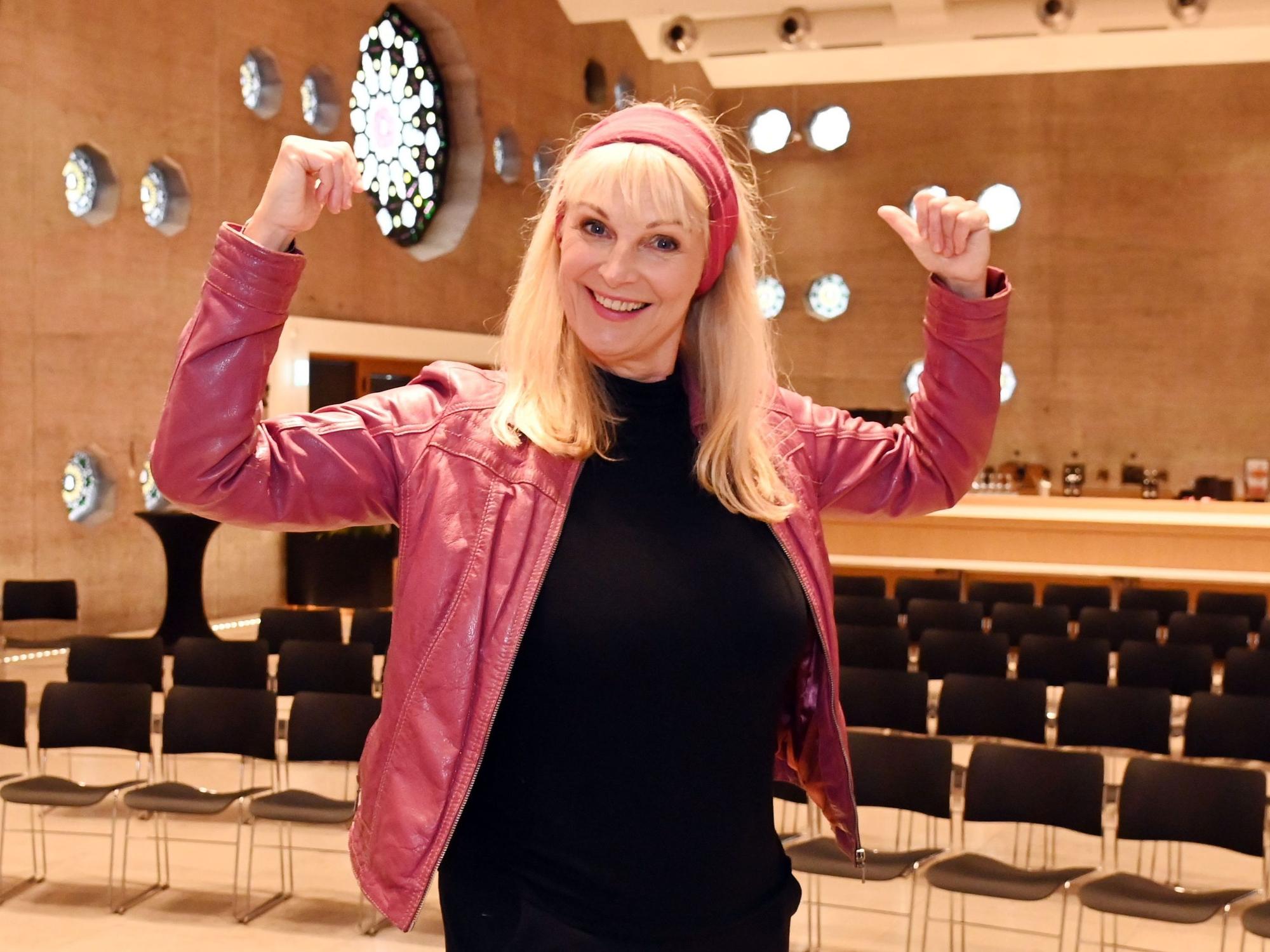 Eine Frau mit roter Lederjacke steht in einem großen Veranstaltungsraum in Siegerpose.
