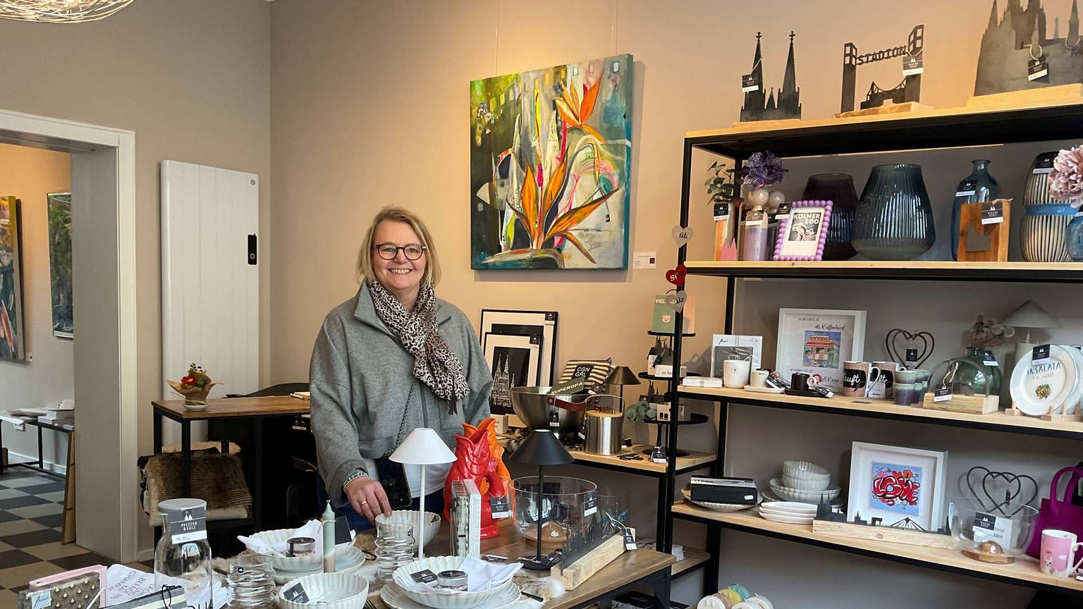Eine Frau mit blondem Haar steht hinter einem Tisch in einem Ladenlokal, in dem allerlei Dekoartikel ausgestellt sind.
