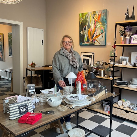 Eine Frau mit blondem Haar steht hinter einem Tisch in einem Ladenlokal, in dem allerlei Dekoartikel ausgestellt sind.
