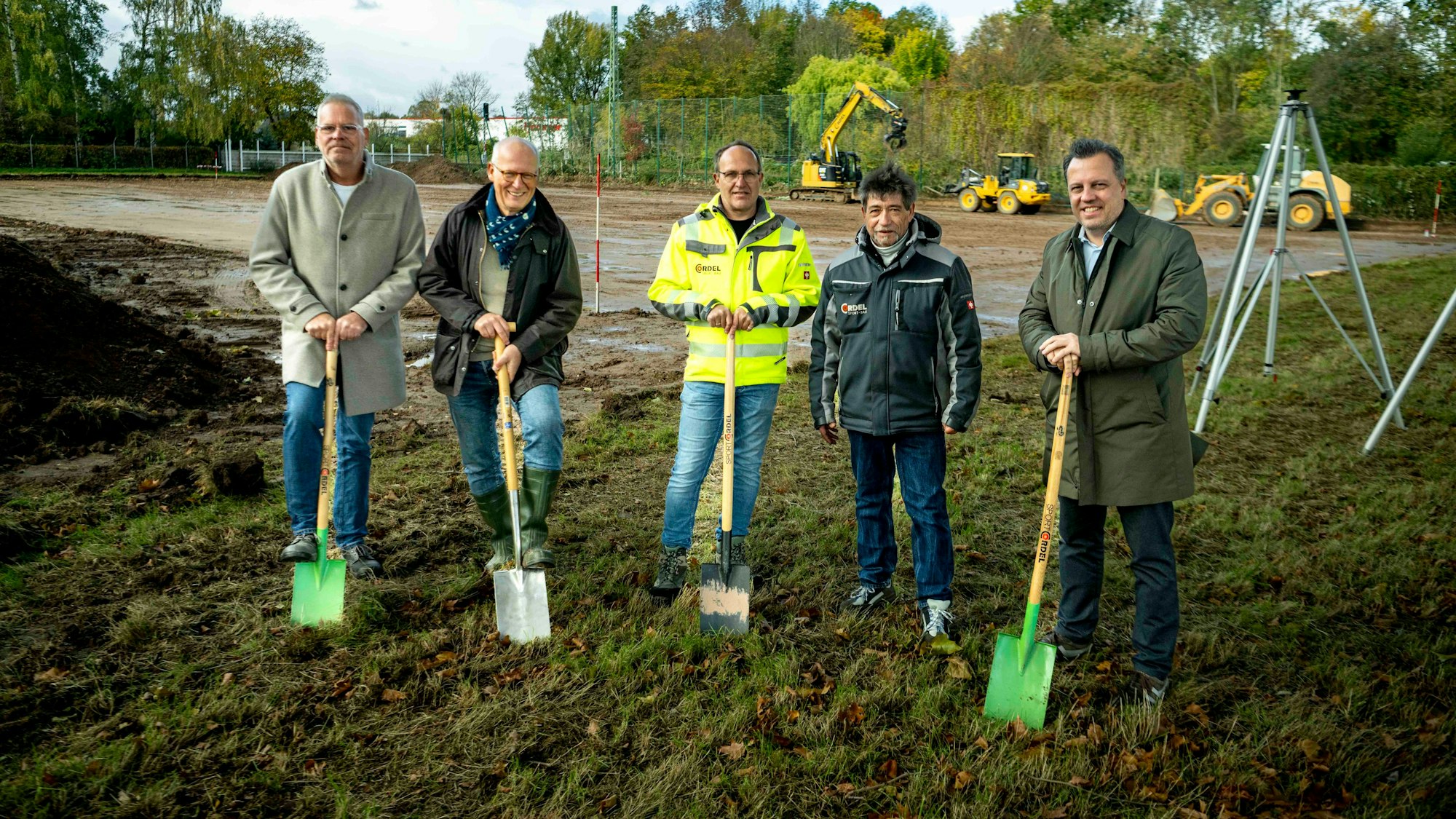 Das Bild zeigt fünf Männer, die einen Spaten halten. Im Hintergrund laufen Bauarbeiten.