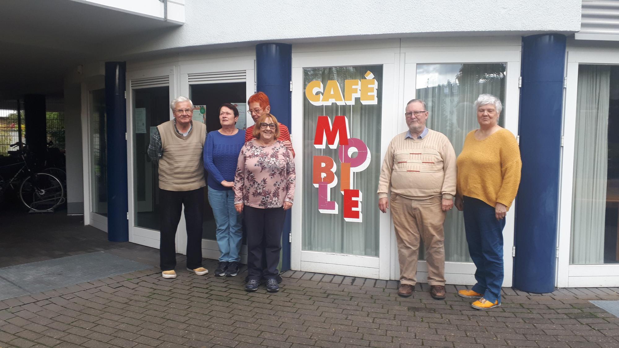 Sechs Senioren stehen linkst und rechts von einem bodentiefen Fenster mit der Aufschrift Café Mobile.