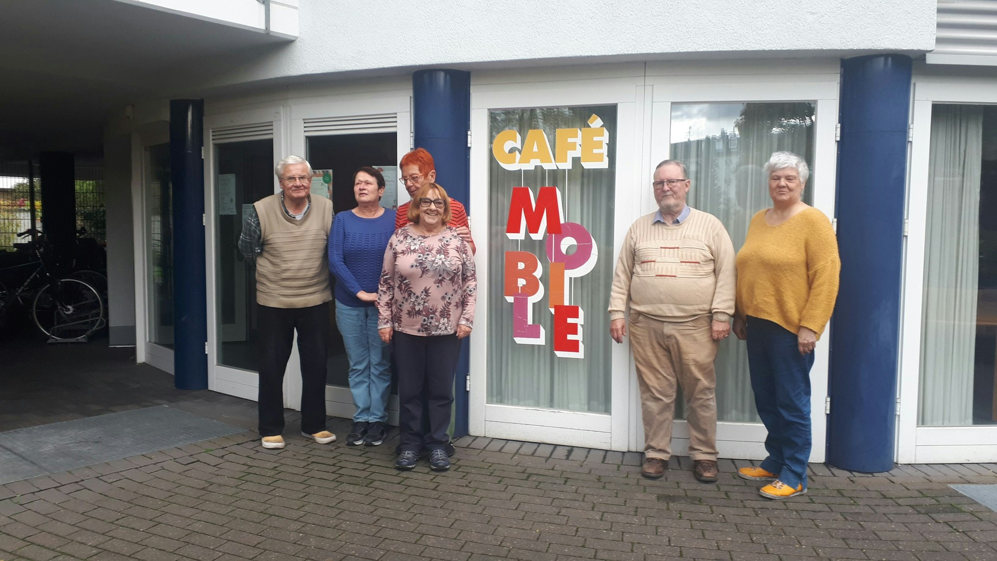 Sechs Senioren stehen linkst und rechts von einem bodentiefen Fenster mit der Aufschrift Café Mobile.