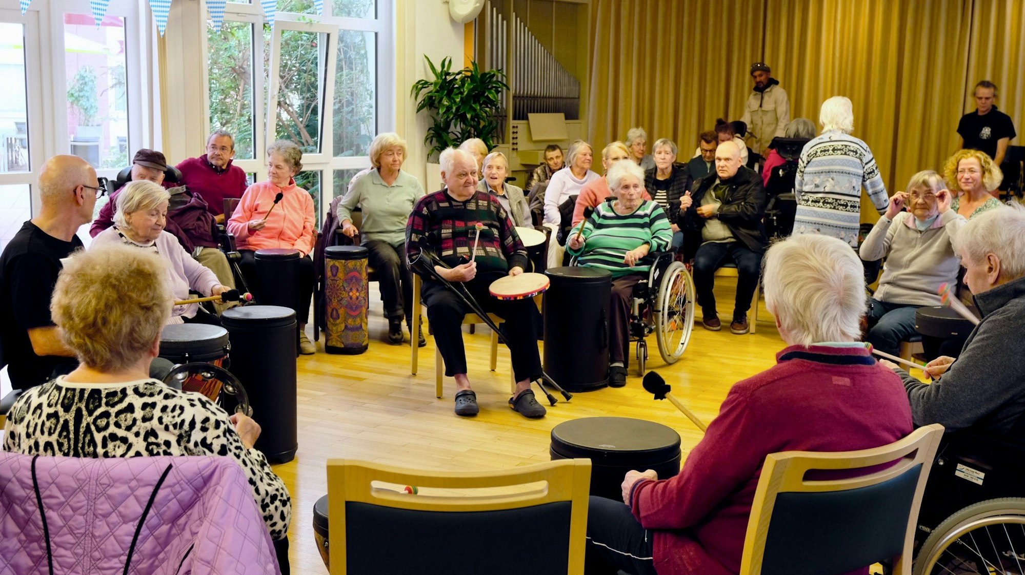 In einem Saal sitzen viele Seniorinnen und Senioren im Kreis, die meisten haben Trommeln vor sich.
