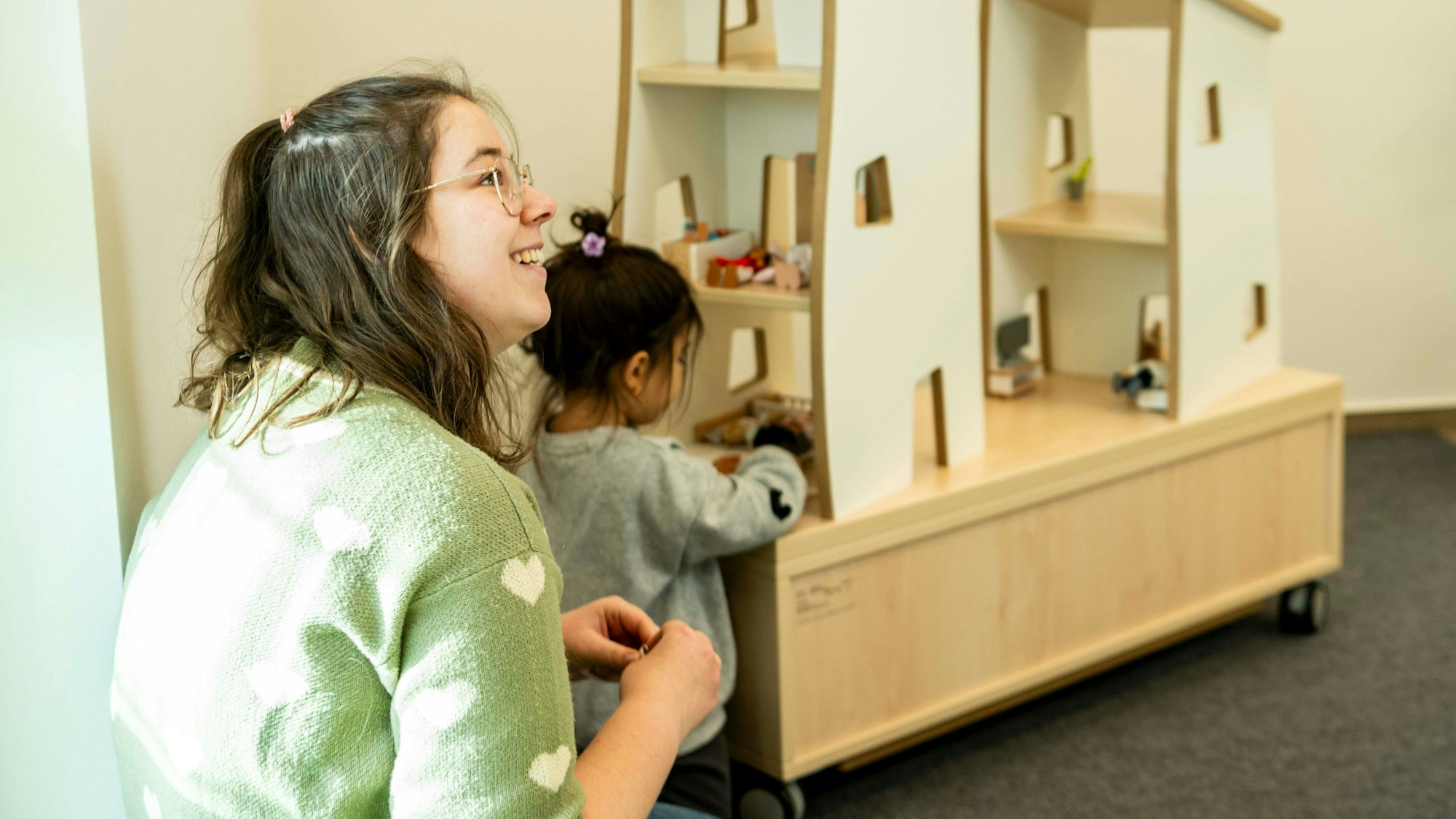 Das Bild zeigt eine lächelnde Erzieherin, die mit einem Kind vor einem modernen Puppenhaus sitzt.