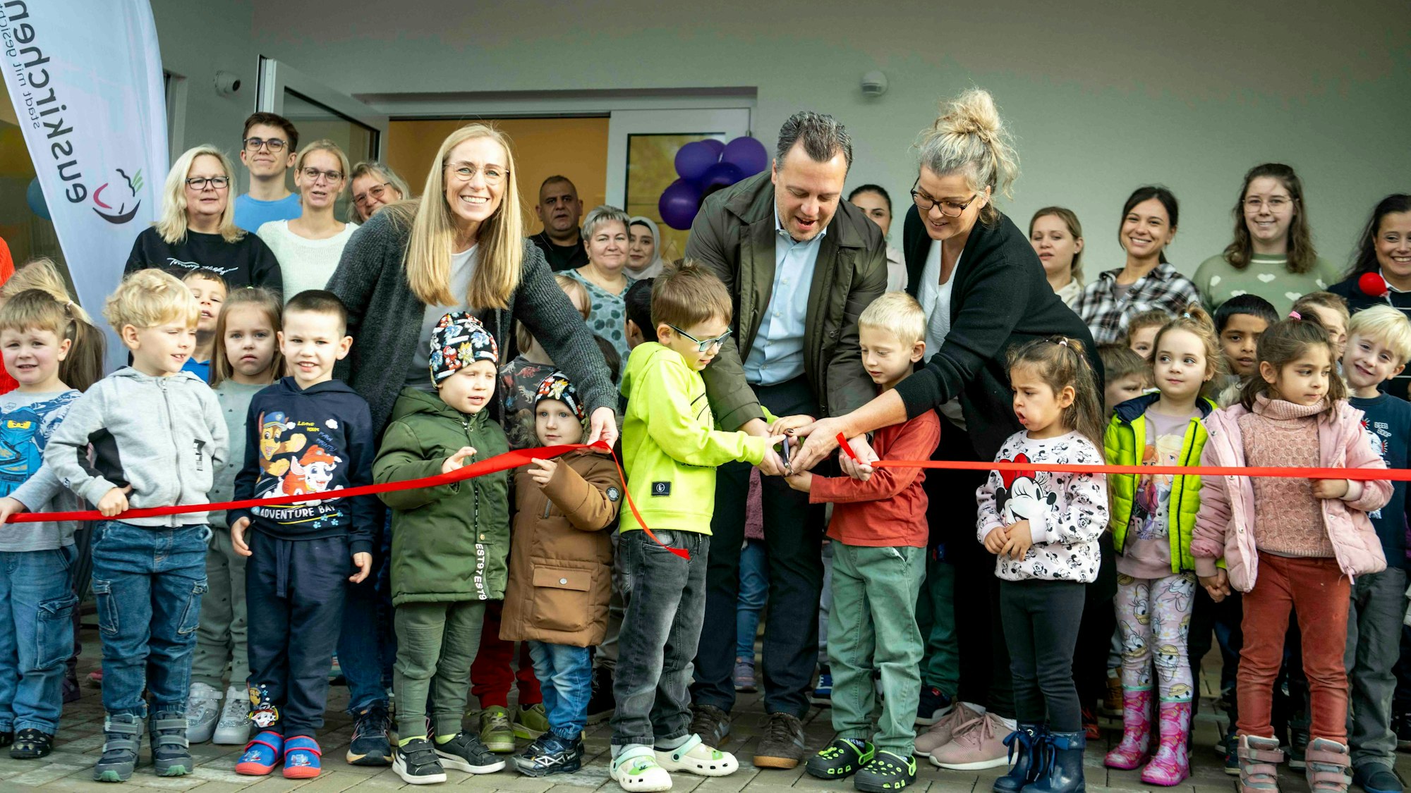 Das Bild zeigt einige Kinder, die mit Bürgermeister Sacha Reichelt und Kita-Leiterin Miriam Zerlett ein rotes Band durchtrennen.