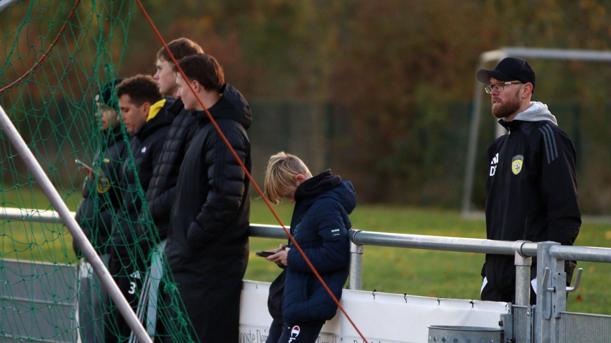 Erftstadt-Lechenichs Trainer Daniel Bartsch steht hinter der Bande, an die sich Zuschauer lehnen, von denen einige aufs Handy schauen.