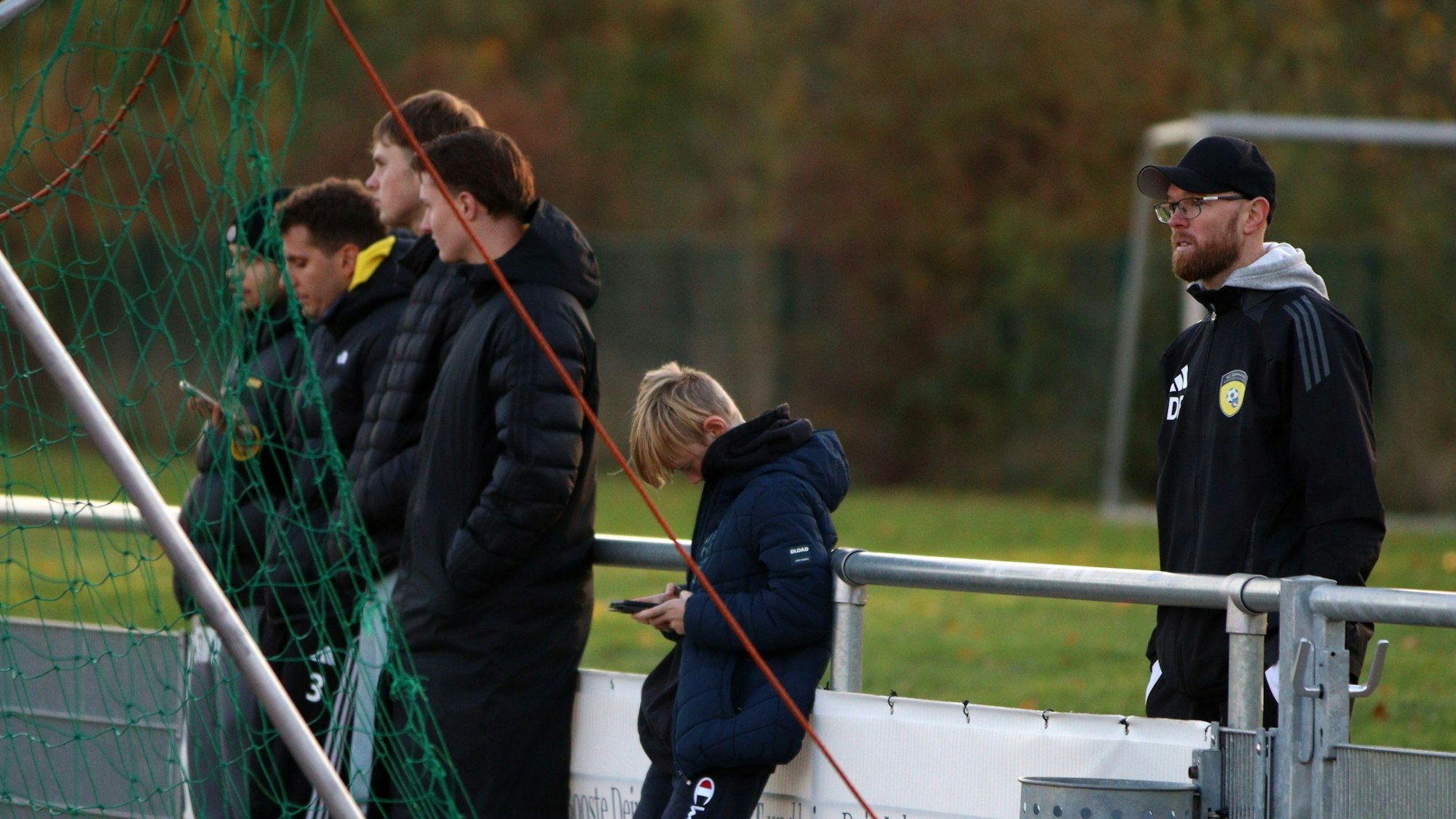 Erftstadt-Lechenichs Trainer Daniel Bartsch steht hinter der Bande, an die sich Zuschauer lehnen, von denen einige aufs Handy schauen.