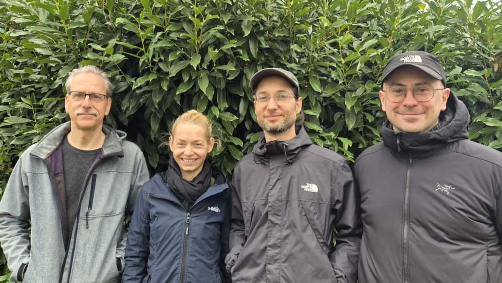Drei Männer und eine Frau stehen nebeneinander vor einer grünen Hecke,