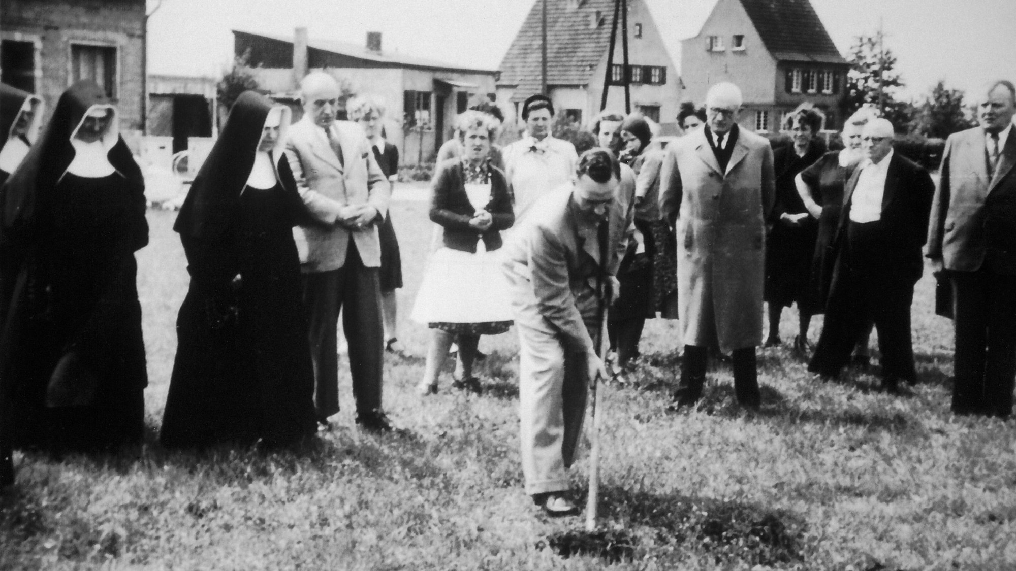 Ein Mann im Anzug führt einen Spatenstich auf einer Wiese aus, daneben drei Nonnen in Ordenstracht.