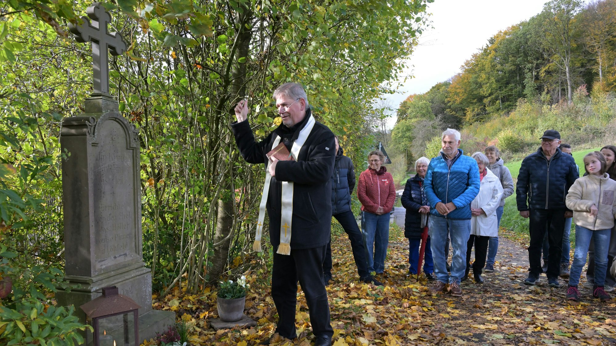 Ein Pfarrer segnet ein Wegekreuz. Dahinter stehen Menschen.
