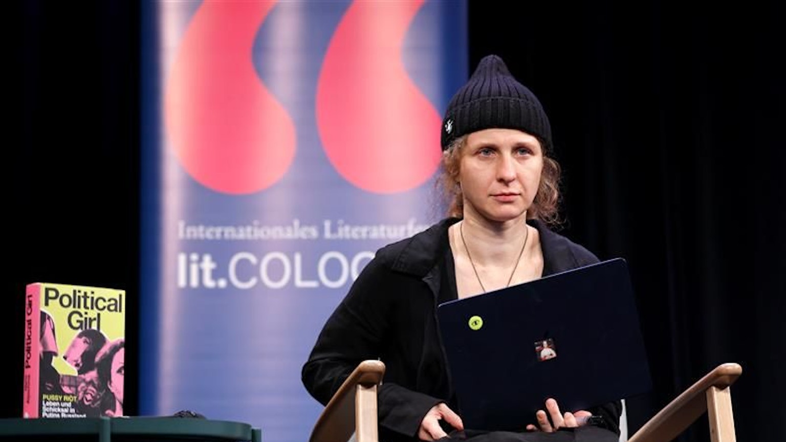 Die Aktivistin trägt schwarz und hat einen Laptop auf dem Schoß. Neben ihr das Buch Political Girl. Im Hintergrund ein Banner der lit.cologne.