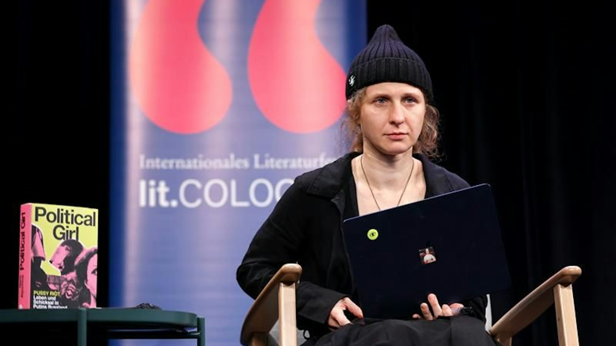 Die Aktivistin trägt schwarz und hat einen Laptop auf dem Schoß. Neben ihr das Buch Political Girl. Im Hintergrund ein Banner der lit.cologne.
