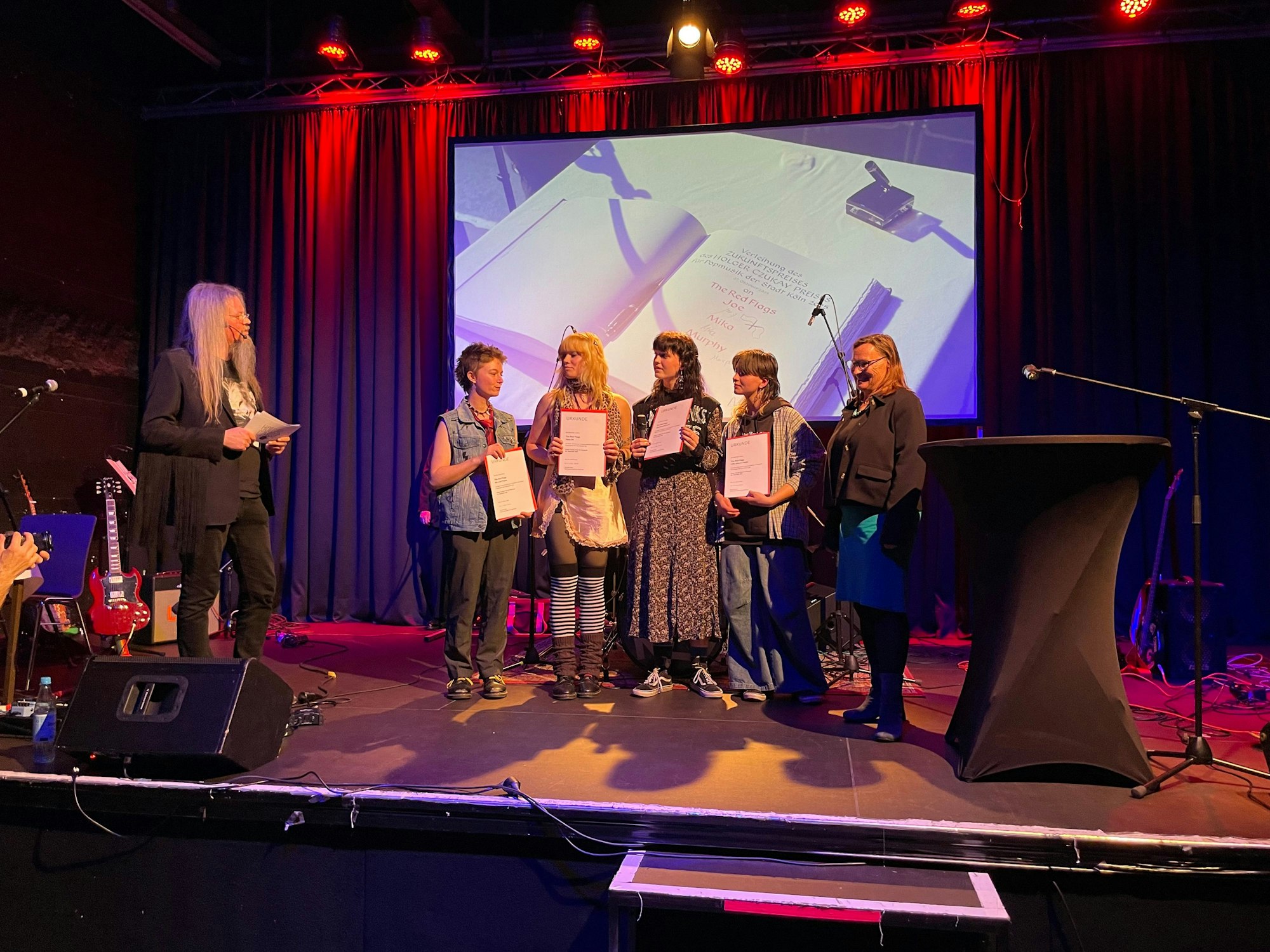 Zu sehen sind vier junge Frauen mit Urkunden in der Hand auf einer Bühne, neben ihnen steht eine Frau und der Moderator.