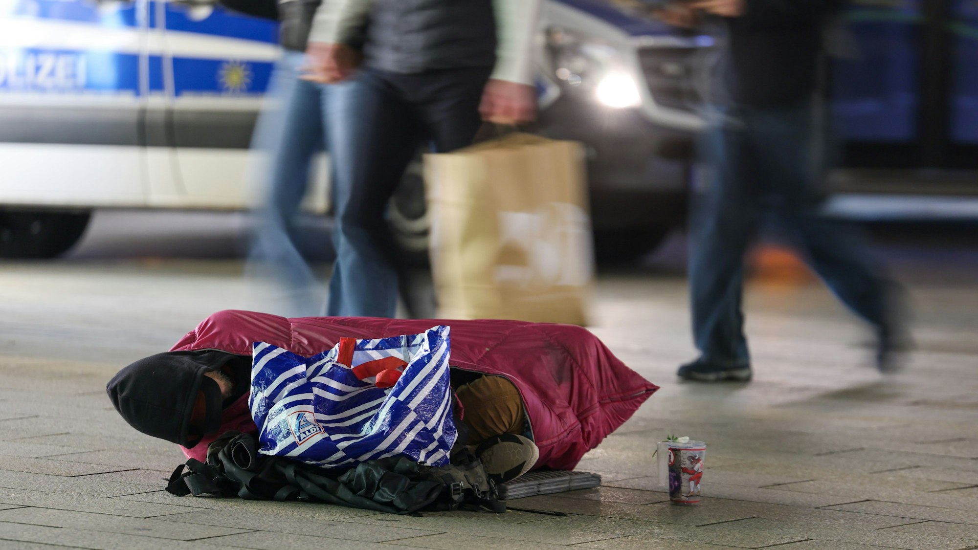 Ein Wohnungsloser liegt in eine Jacke gehüllt in einer Fußgängerzone während Passanten vorbeigehen.