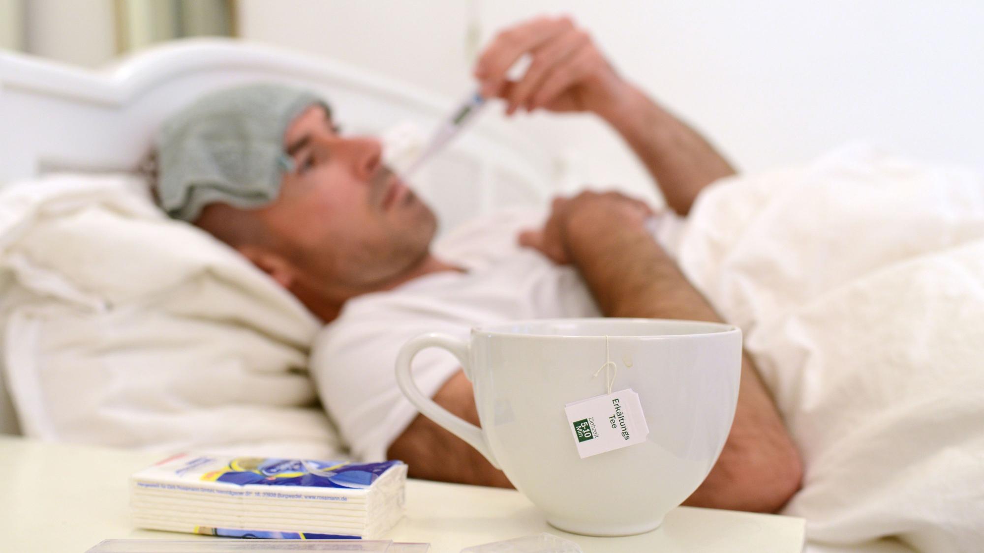 Ein kranker Mann liegt im Bett und misst Fieber, auf dem Tisch vor ihm steht eine Tasse mit Erkältungstee.