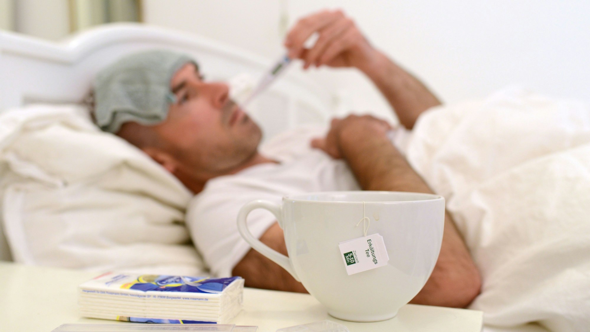 Ein kranker Mann liegt im Bett und misst Fieber, auf dem Tisch vor ihm steht eine Tasse mit Erkältungstee.
