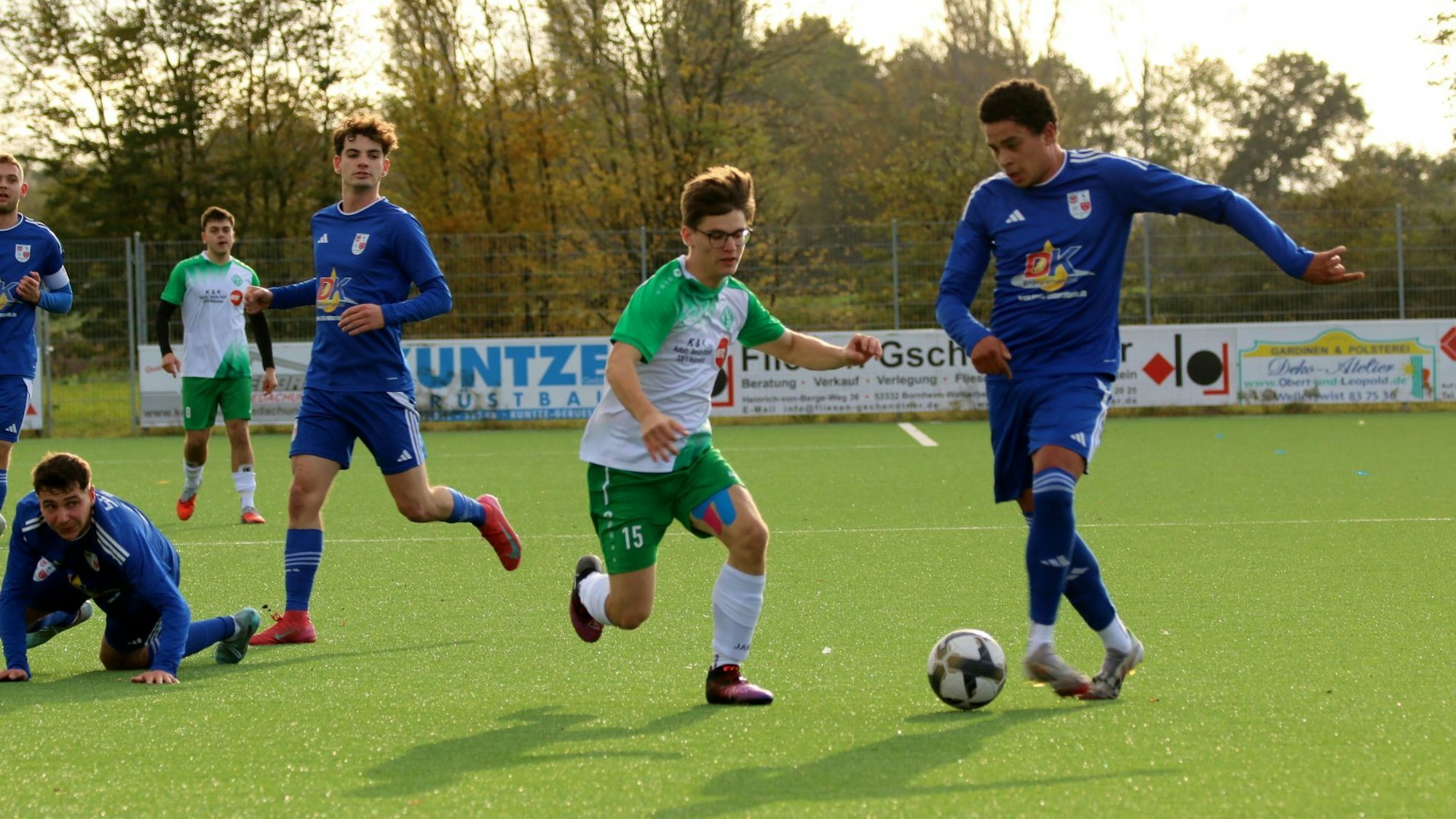 Zwei Gegenspieler rennen auf einen Fußball zu. Mehrere Mitspieler schauen in Richtung des Zweikampfs.