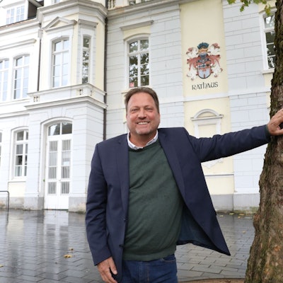 Ein Mann im blauen Sakko neben einem Baum, im Hintergrund an einer Fassade das Wappen der Stadt Königswinter und der Schriftzug Rathaus.